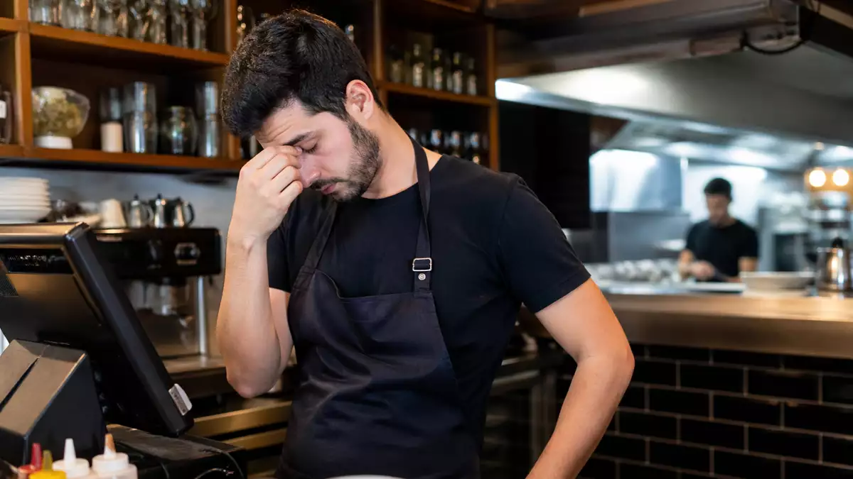 Man in a dark apron at a cafe counter rubs his eyes in frustration while working.