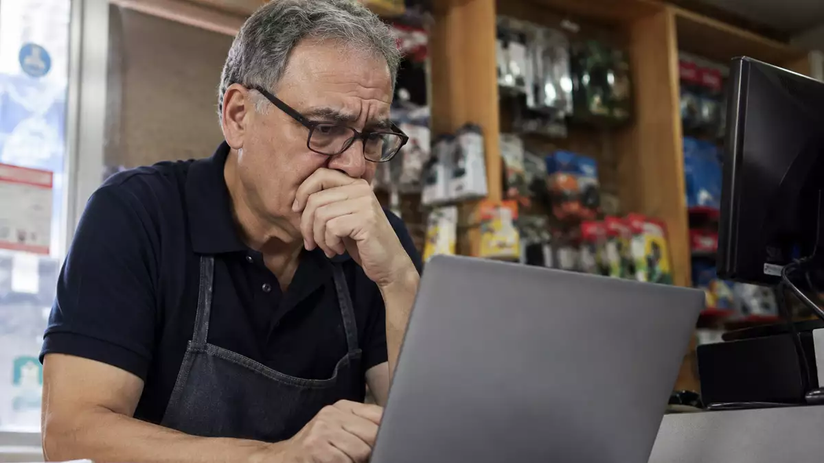 Older man in a shop wearing a dark polo and apron, focused on a laptop at the counter, hand covering his mouth in thought.