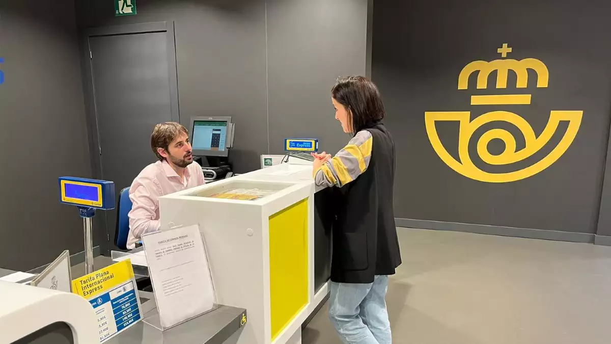 Empleado de Correos atendiendo en oficina mientras una clienta realiza una gestión en ventanilla