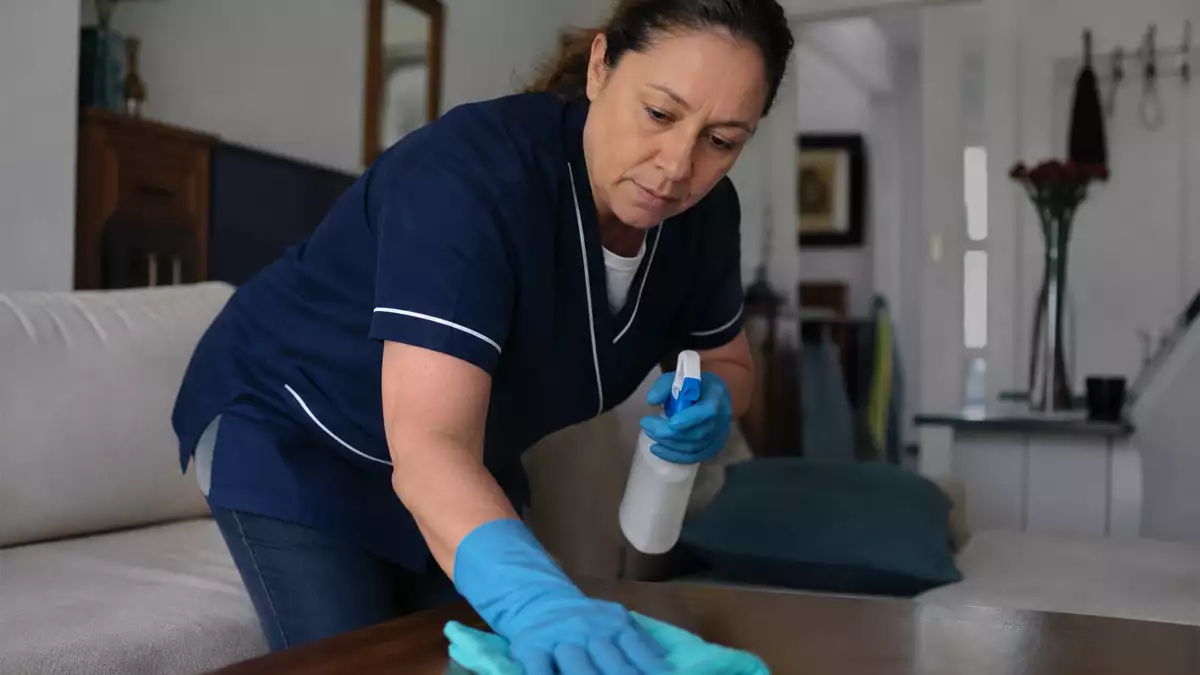 Caregiver wearing blue uniform and gloves spraying cleaner on a wooden table in a living room