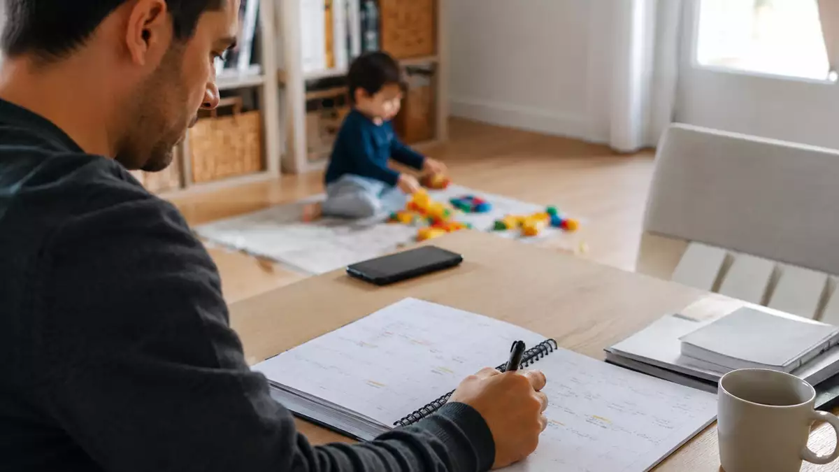 Man writing in a notebook at a desk with a coffee mug nearby while a child plays with colorful blocks in the background.