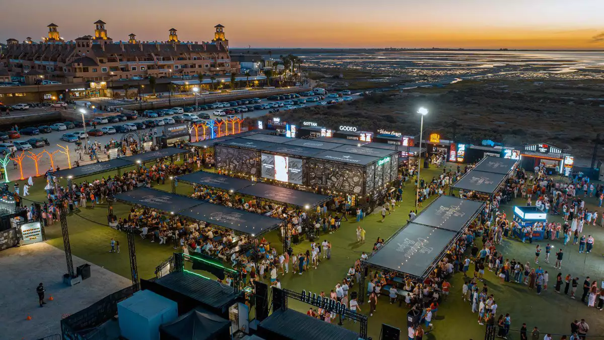 Outdoor coastal festival at sunset with multiple stages, long dining tables, and crowds enjoying the event by the sea.
