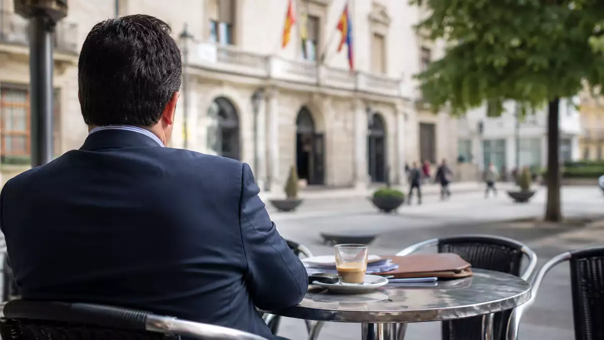 Funcionario sentado en la terraza de un bar durante la mañana frente a un ayuntamiento mientras aparenta estar en su jornada laboral