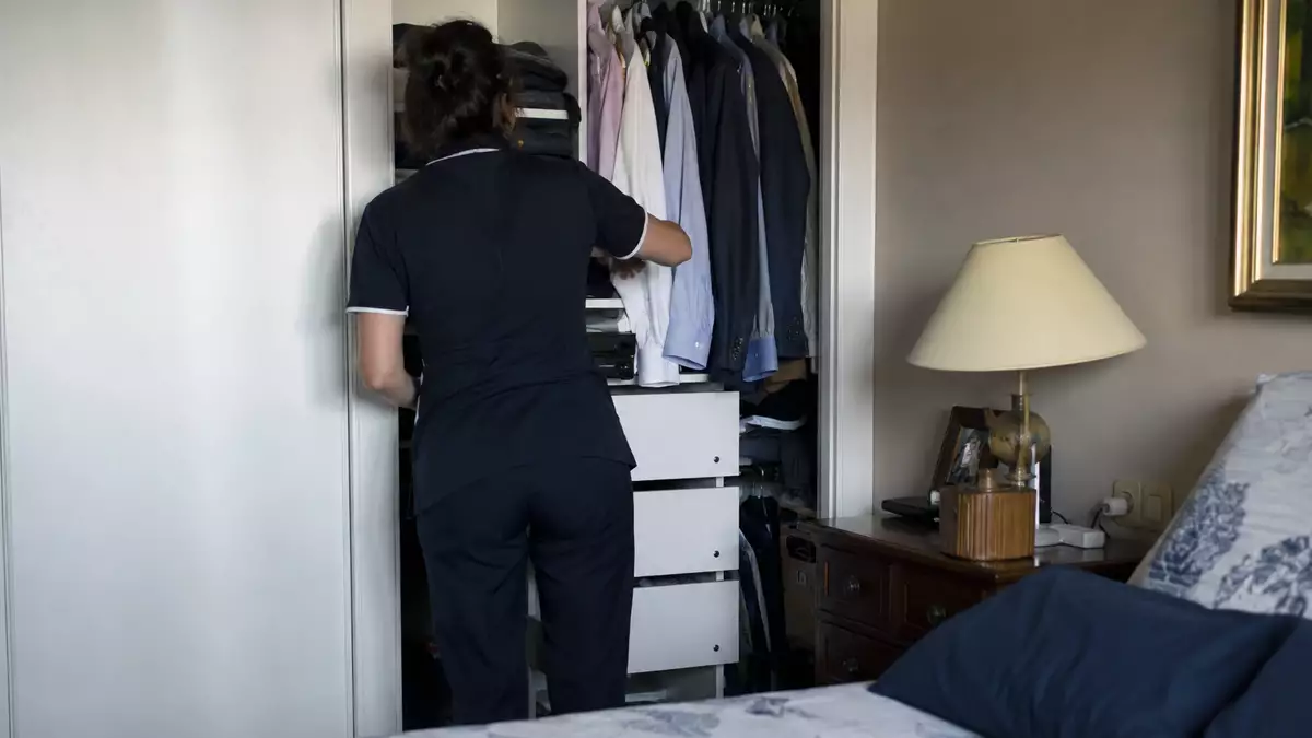 Care worker in a navy uniform organizing clothes in an open bedroom closet, with a dresser and lamp nearby