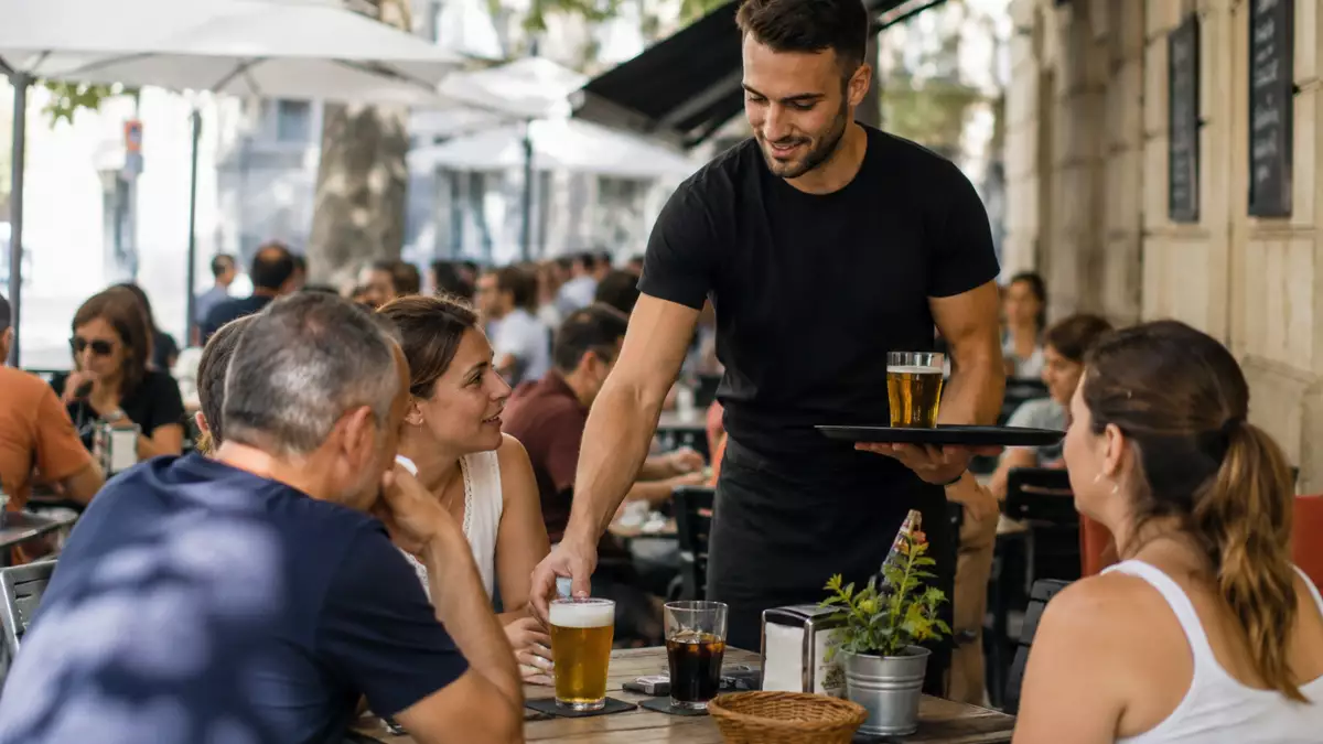 Camarero sirviendo bebidas a varios clientes en la terraza de un restaurante durante la jornada laboral.