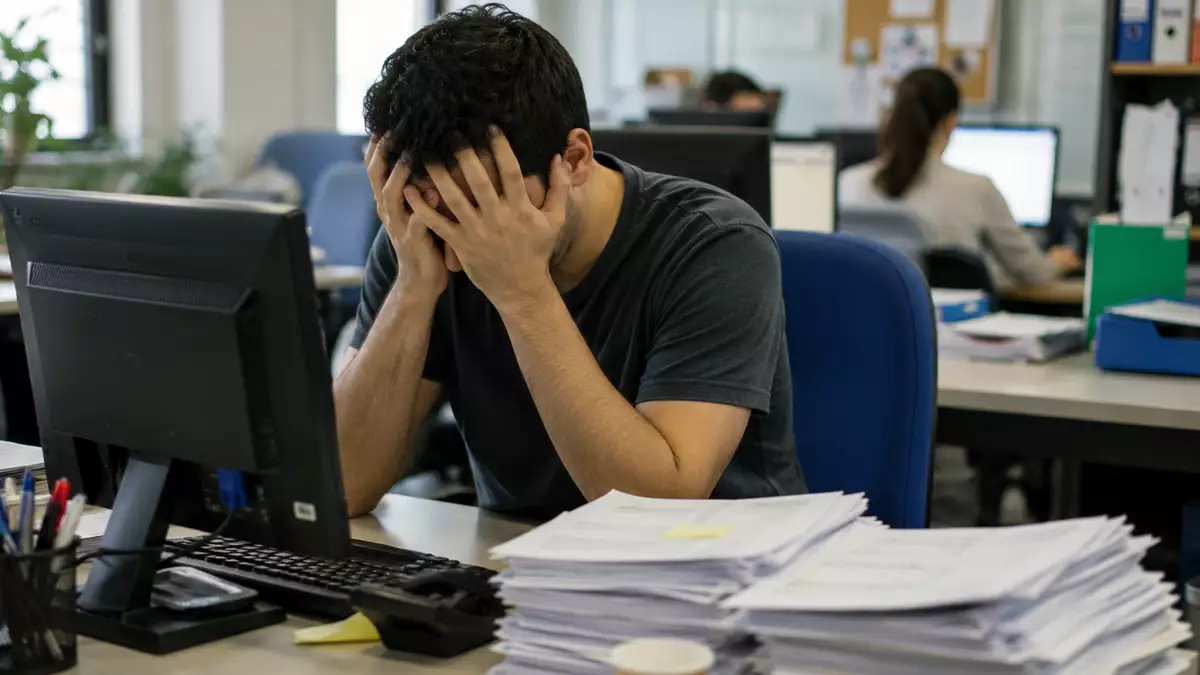 Overwhelmed office worker covering his face at a desk with a computer and a tall stack of papers.