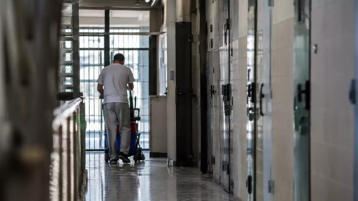 A man in light clothing pushes a cleaning cart down a prison hallway lined with cell doors and bars at the end of the corridor.